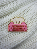 A pink and gold enamel pin shaped like a tissue box with the text 'CRYING OVER FICTIONAL CHARACTERS' on a white knit background.