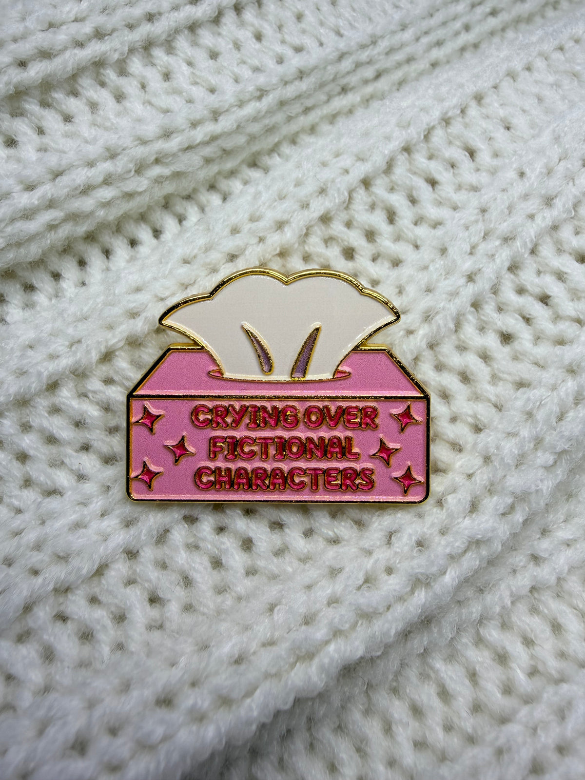 A pink and gold enamel pin shaped like a tissue box with the text 'CRYING OVER FICTIONAL CHARACTERS' on a white knit background.