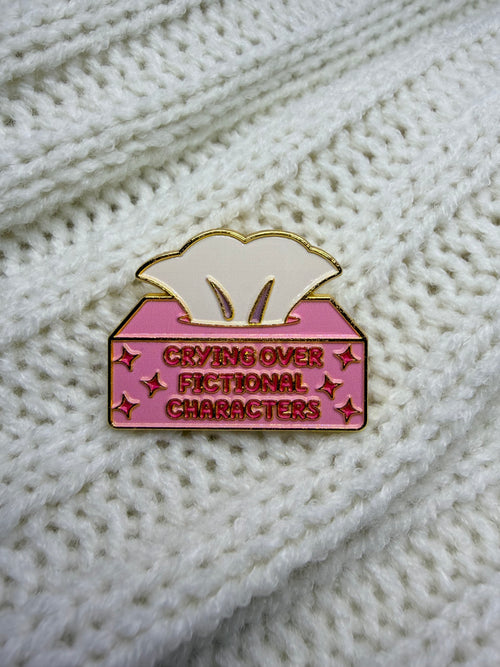 A pink and gold enamel pin shaped like a tissue box with the text 'CRYING OVER FICTIONAL CHARACTERS' on a white knit background.