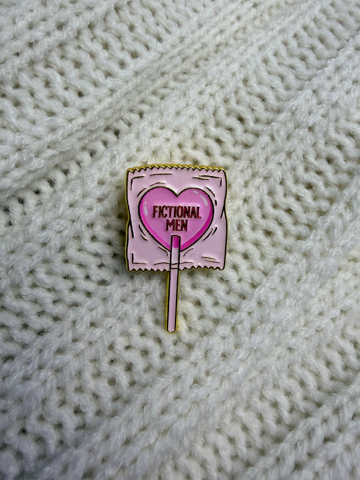 A small enamel pin shaped like a pink heart on a stick, placed on a cream-colored knitted fabric background. The pin reads 'FICTIONAL MEN' in black text.