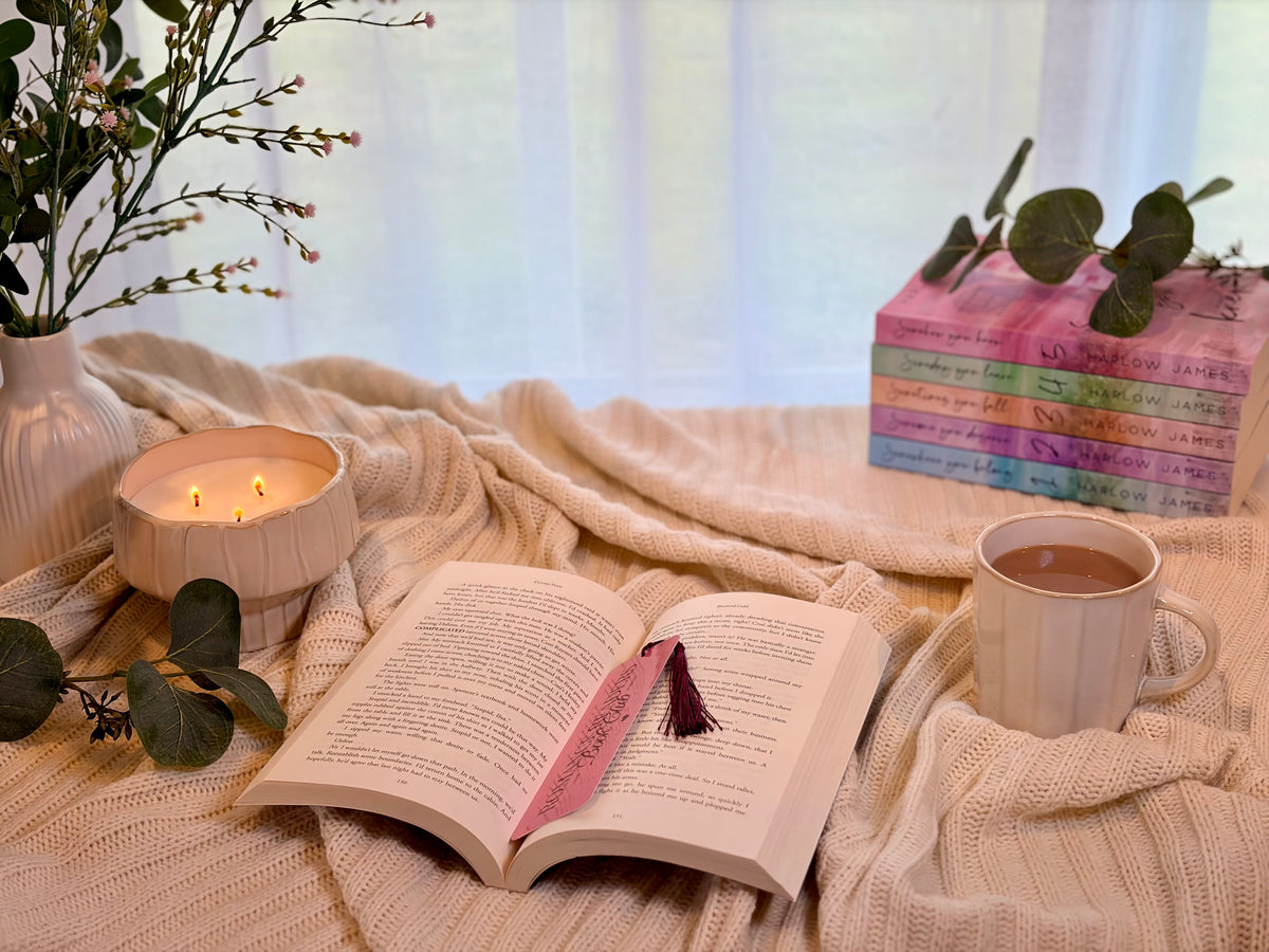 A cozy lifestyle scene featuring an open book with a pink tassel bookmark on a cream knit blanket, next to a lit candle and a cup of tea, with a stack of colorful books and a vase of flowers in the background.