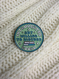 A round enamel pin with the text 'INTROVERTED BUT WILLING TO DISCUSS BOOKS' in white and blue lettering on a mint green background, placed on a cream-colored knitted sweater.