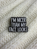 A black and white enamel pin with the text 'I'M NICER THAN MY FACE LOOKS' resting on a cream-colored knitted blanket.