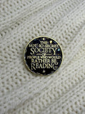 A round enamel pin with a gold border and black background featuring the text 'THE NOT SO-SECRET SOCIETY OF PEOPLE WHO WOULD RATHER BE READING' in gold lettering, displayed on a cream-colored knitted sweater.