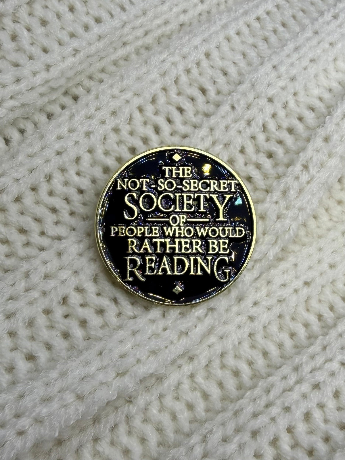 A round enamel pin with a gold border and black background featuring the text 'THE NOT SO-SECRET SOCIETY OF PEOPLE WHO WOULD RATHER BE READING' in gold lettering, displayed on a cream-colored knitted sweater.