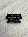 A close-up of a black enamel pin with white text reading 'I work to support my reading addiction' placed on a cream-colored knitted fabric background.
