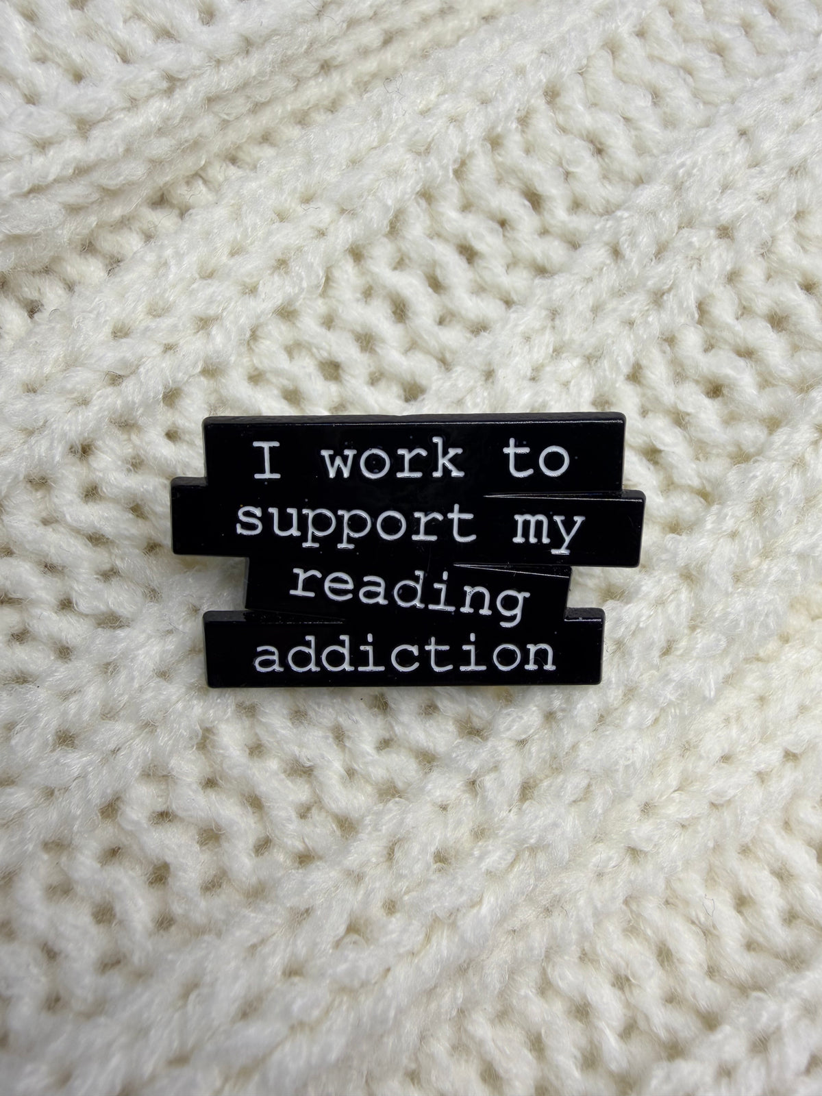 A close-up of a black enamel pin with white text reading 'I work to support my reading addiction' placed on a cream-colored knitted fabric background.
