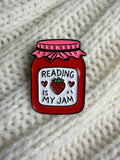 A red enamel pin shaped like a jar of jam with a pink lid and a white label that reads 'READING IS MY JAM' with a strawberry in the center, placed on a cream-colored knitted blanket.