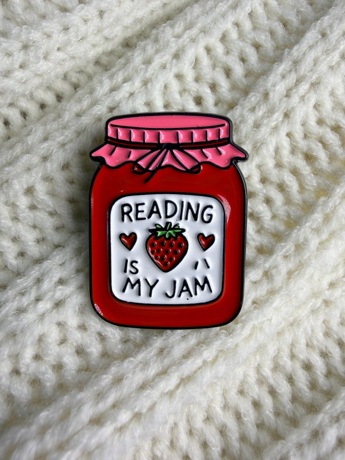 A red enamel pin shaped like a jar of jam with a pink lid and a white label that reads 'READING IS MY JAM' with a strawberry in the center, placed on a cream-colored knitted blanket.