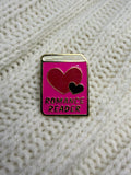 A rectangular enamel pin shaped like a book with a bright pink cover, featuring a red heart, a smaller black heart, and the words 'ROMANCE READER' in black lettering. The pin is placed on a textured cream-colored knit fabric background.