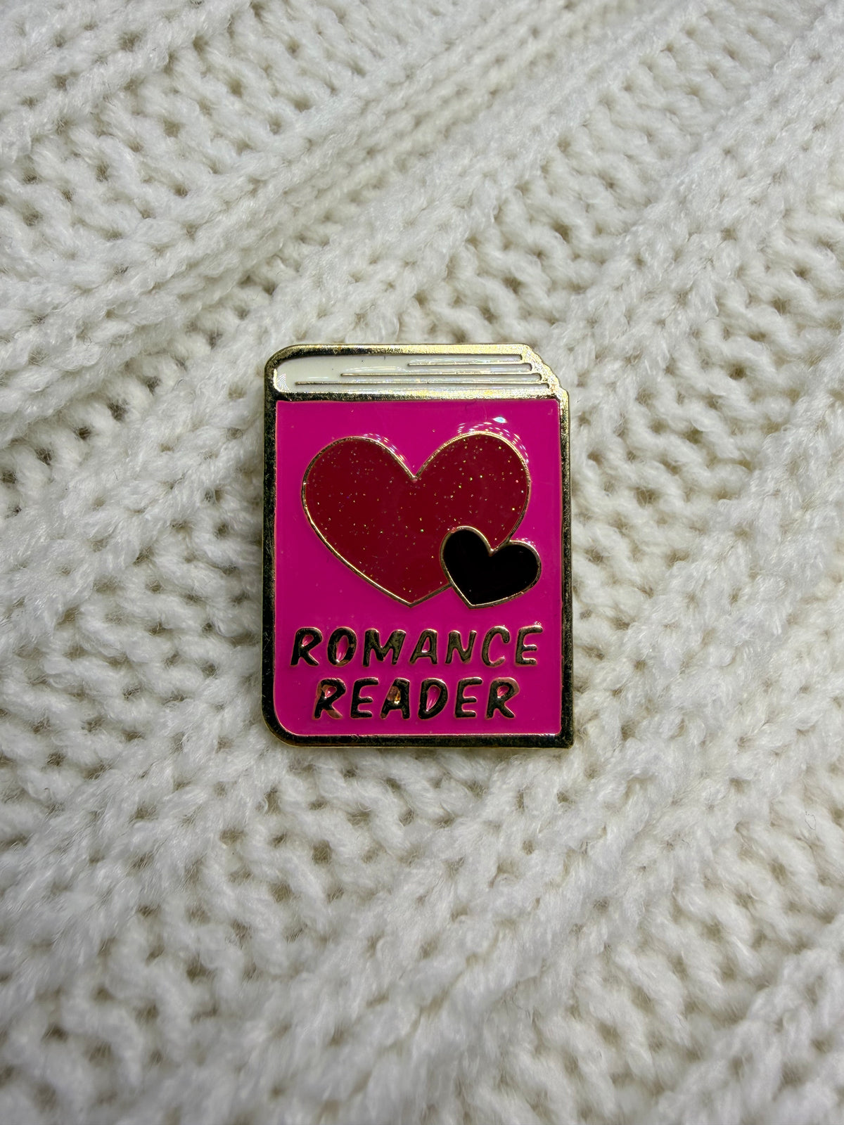 A rectangular enamel pin shaped like a book with a bright pink cover, featuring a red heart, a smaller black heart, and the words 'ROMANCE READER' in black lettering. The pin is placed on a textured cream-colored knit fabric background.