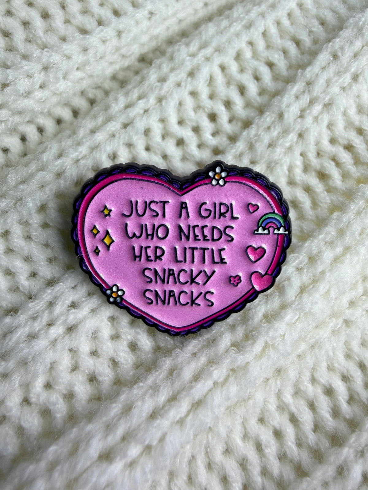 A heart-shaped enamel pin with the text 'JUST A GIRL WHO NEEDS HER LITTLE SNACKY SNACKS' in black lettering on a pink background, decorated with small flowers, stars, and a rainbow. The pin is displayed on a textured cream-colored knit blanket.